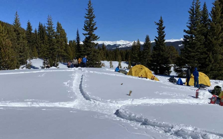 yellow tents are set up in a snowy clearing amongst trees. There are dug out walking paths throughout the campsite and mountains in the background. 